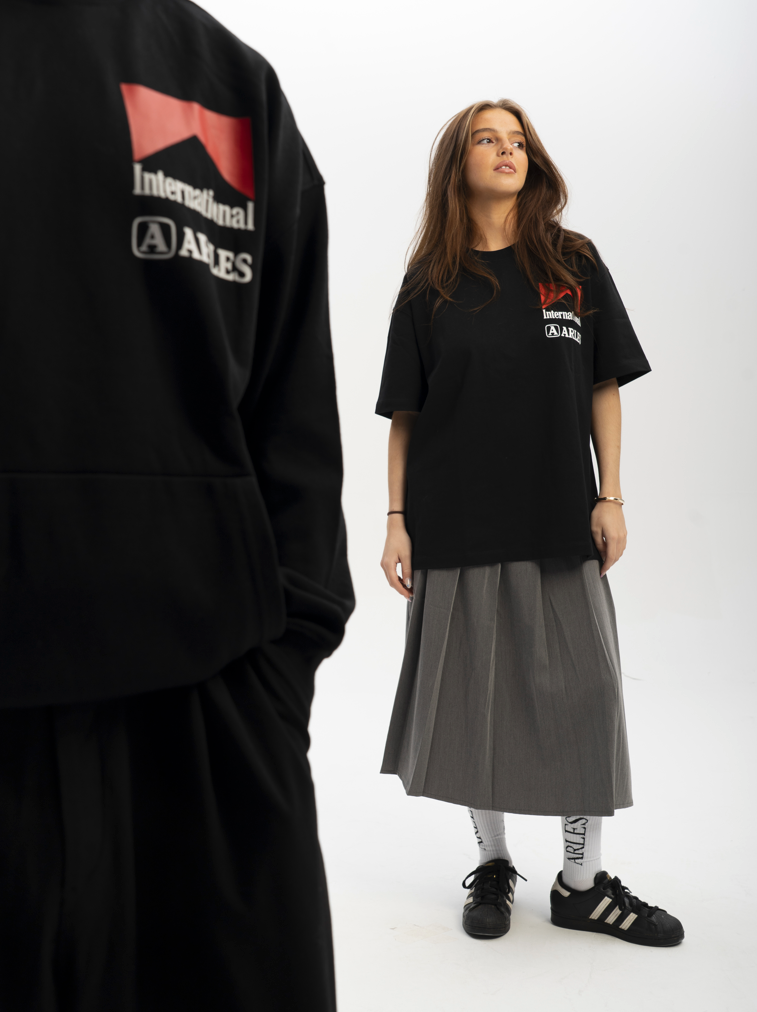 Person wearing a black t-shirt with a logo and gray skirt on a white background ARLES